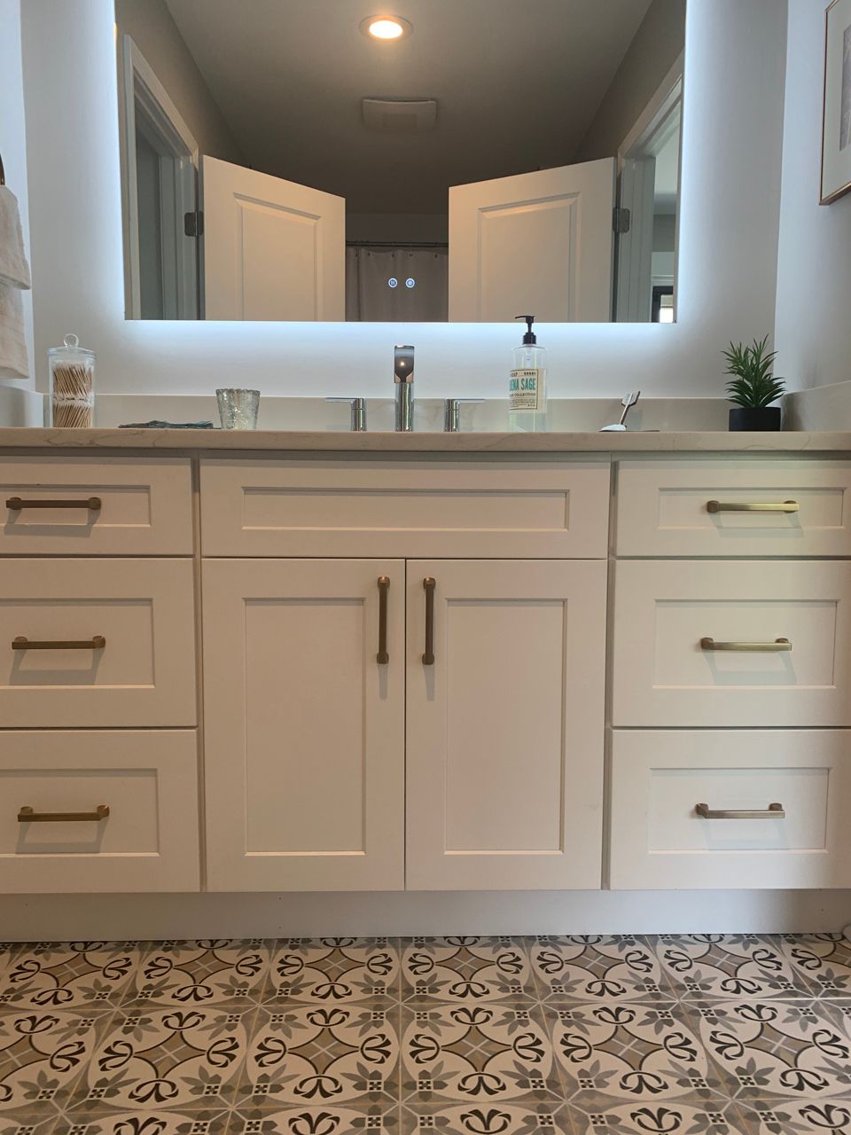 Bathroom vanity with white cabinets, gray countertop, illuminated mirror, and patterned floor.