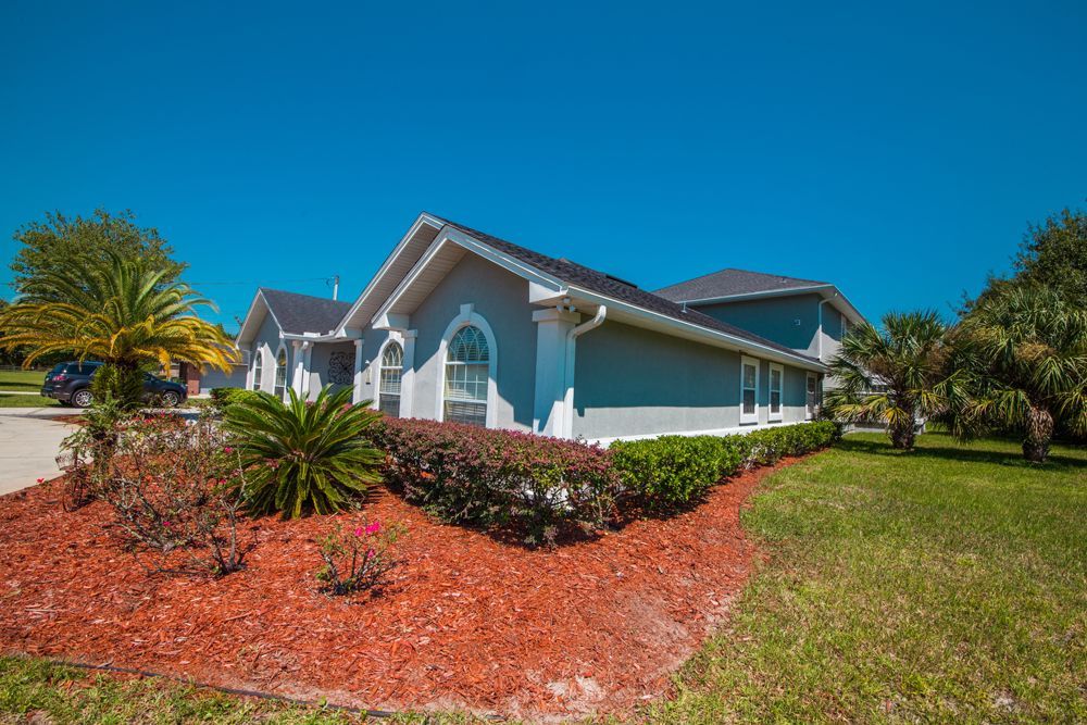 A light blue house with a dark roof, surrounded by red mulch, green grass, and sunny sky.