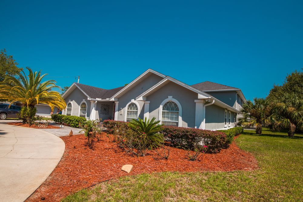 Gray house with red mulch, palm trees, and green grass under a clear blue sky.