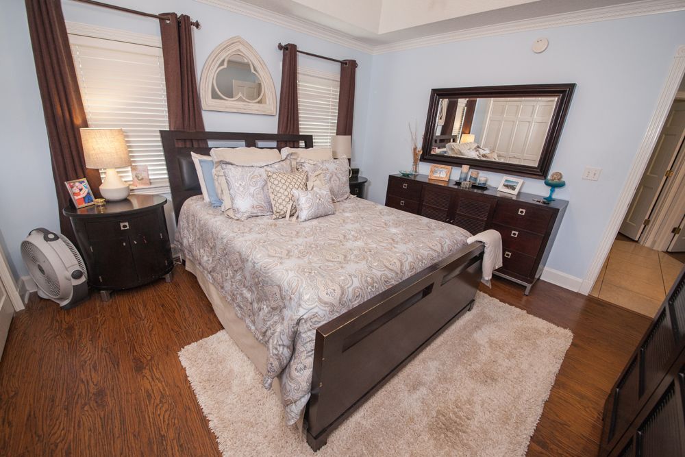 Bedroom with bed, dresser, nightstand, and fan. Light blue walls, brown furniture, and wooden floors.
