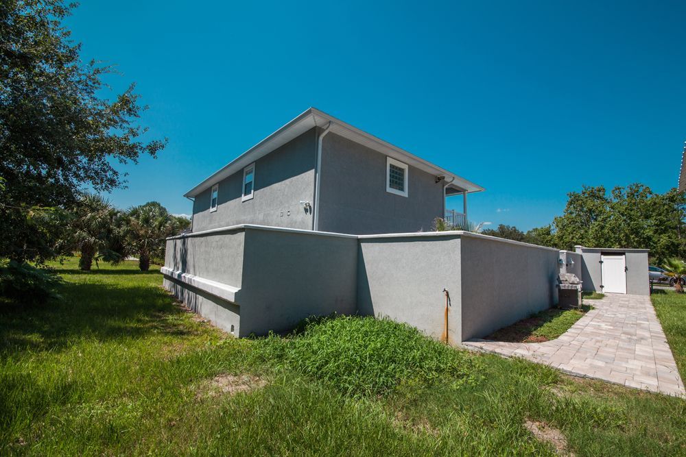 Two-story gray house with a wall, sidewalk, and overgrown grass on a sunny day.