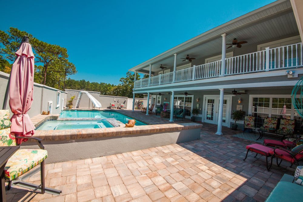 Backyard patio with pool, two-story house with balconies, brick pavers, and outdoor furniture under a blue sky.