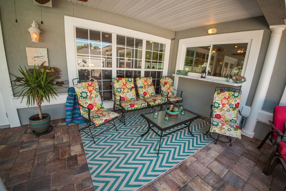 Patio with outdoor seating, rug, and plants. Colorful floral chairs, teal rug, brick floor.