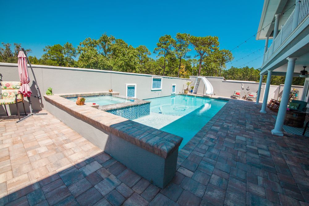 Backyard pool with spa, slide, and paved patio under a blue sky.