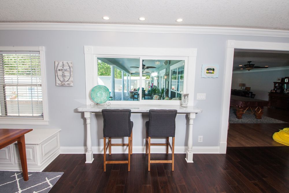 Two gray bar stools face a long white table with a large window overlooking a sunroom.