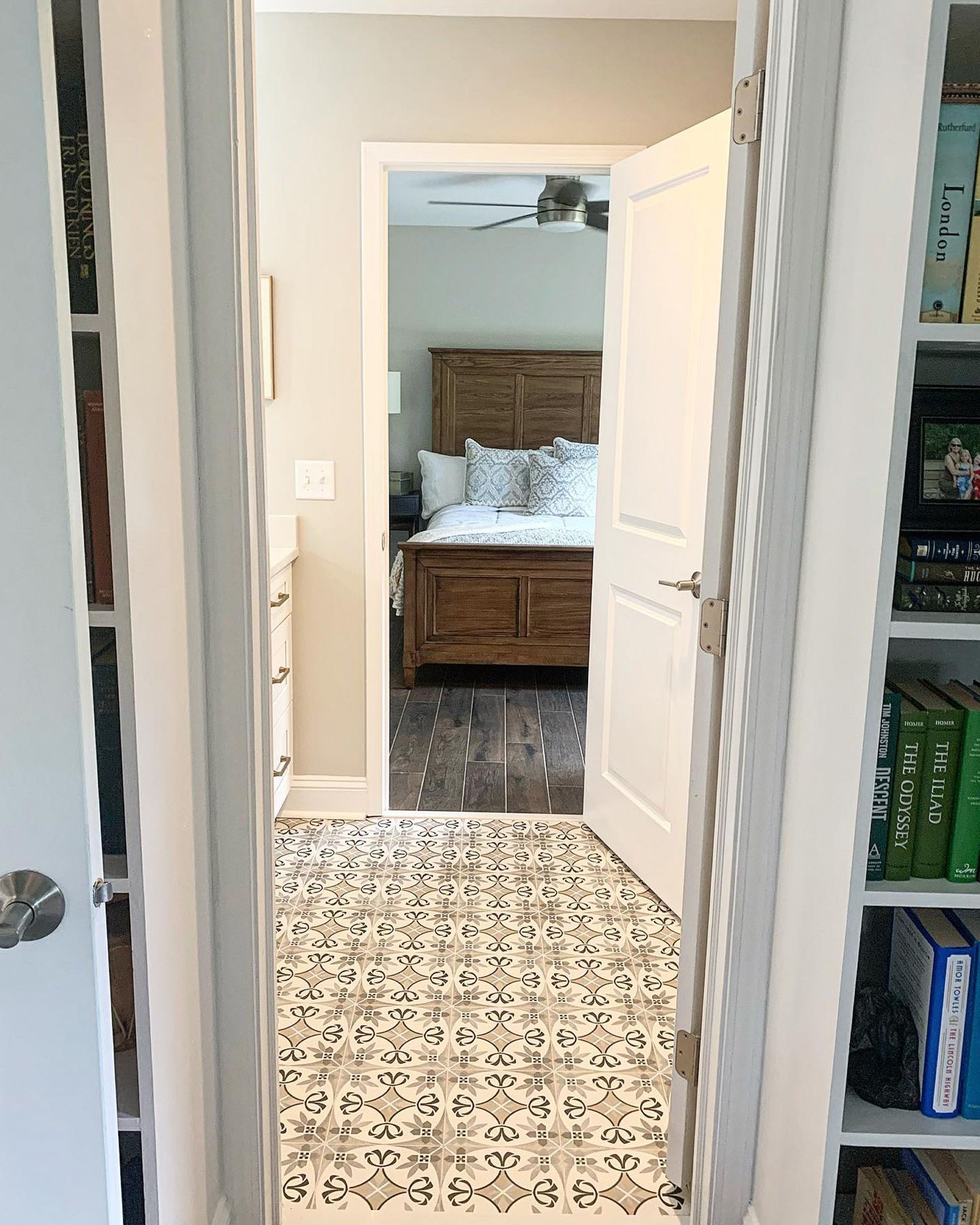 A view through an open doorway into a bedroom, revealing a patterned floor, bed, and bookshelves.