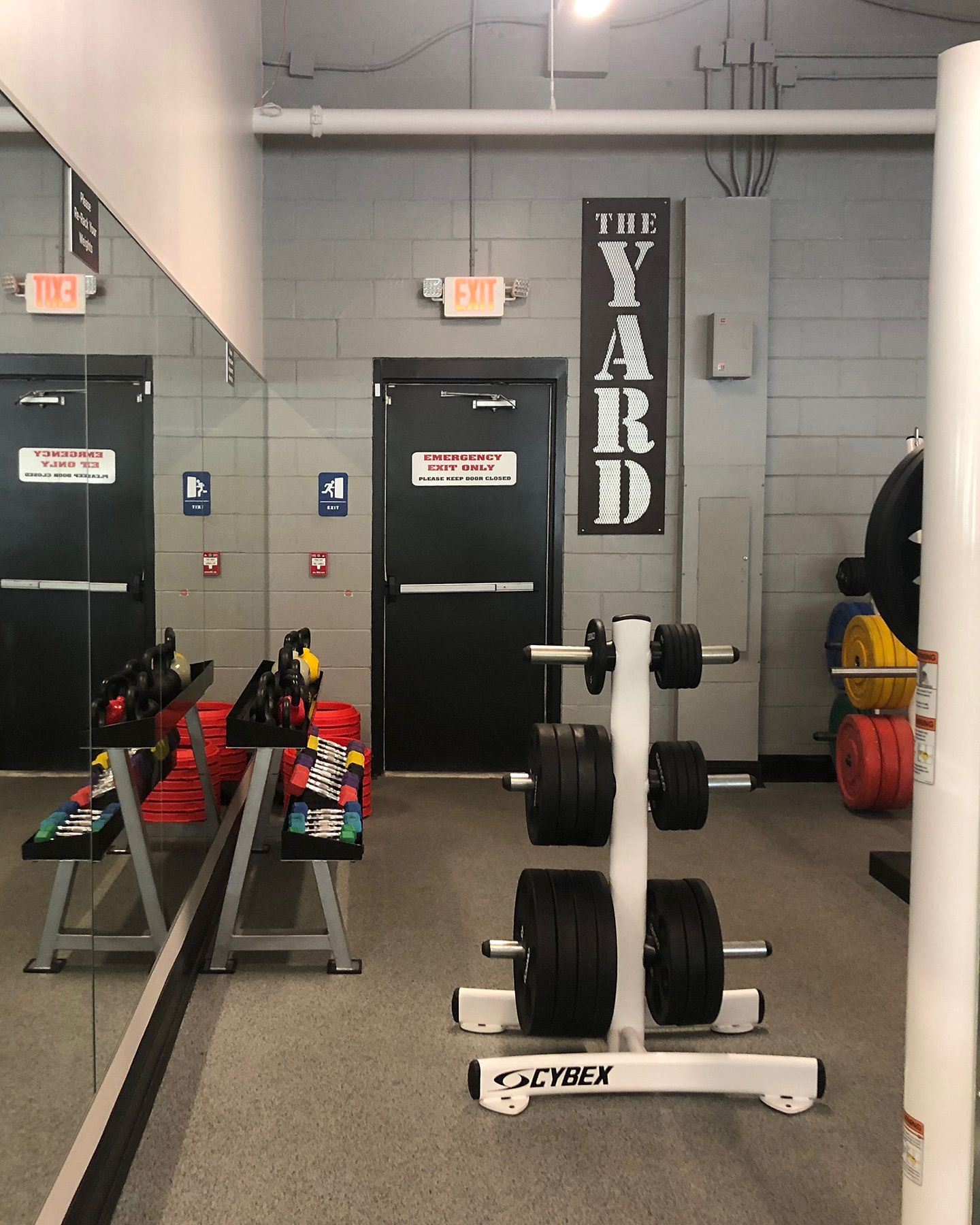 Weight room with weights, benches, mirrors, and exit doors. Sign reads