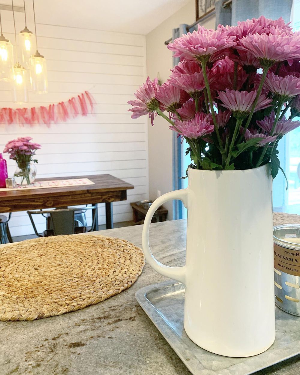 White pitcher overflowing with pink flowers on a countertop near a wooden table.