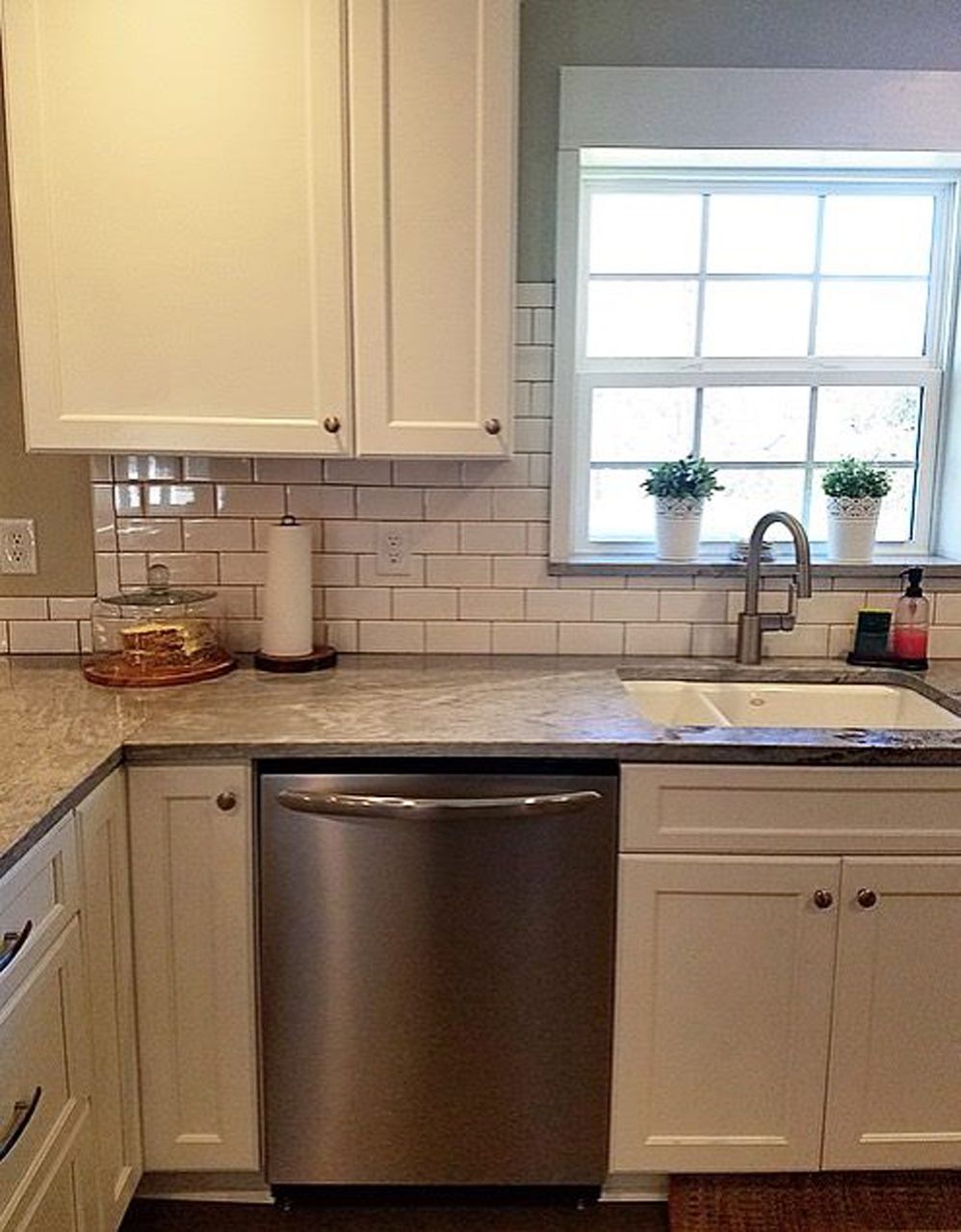 Kitchen corner with white cabinets, stainless steel dishwasher, sink, and window with plants.