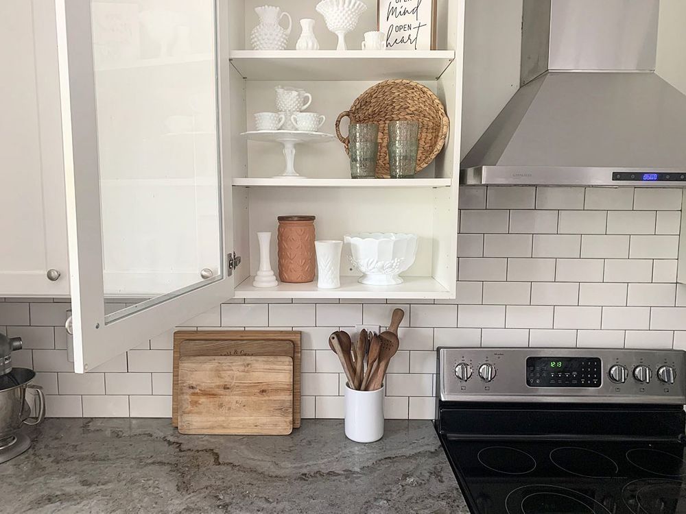 White kitchen cabinet with open door, holding white dishes and decor, above a stove.