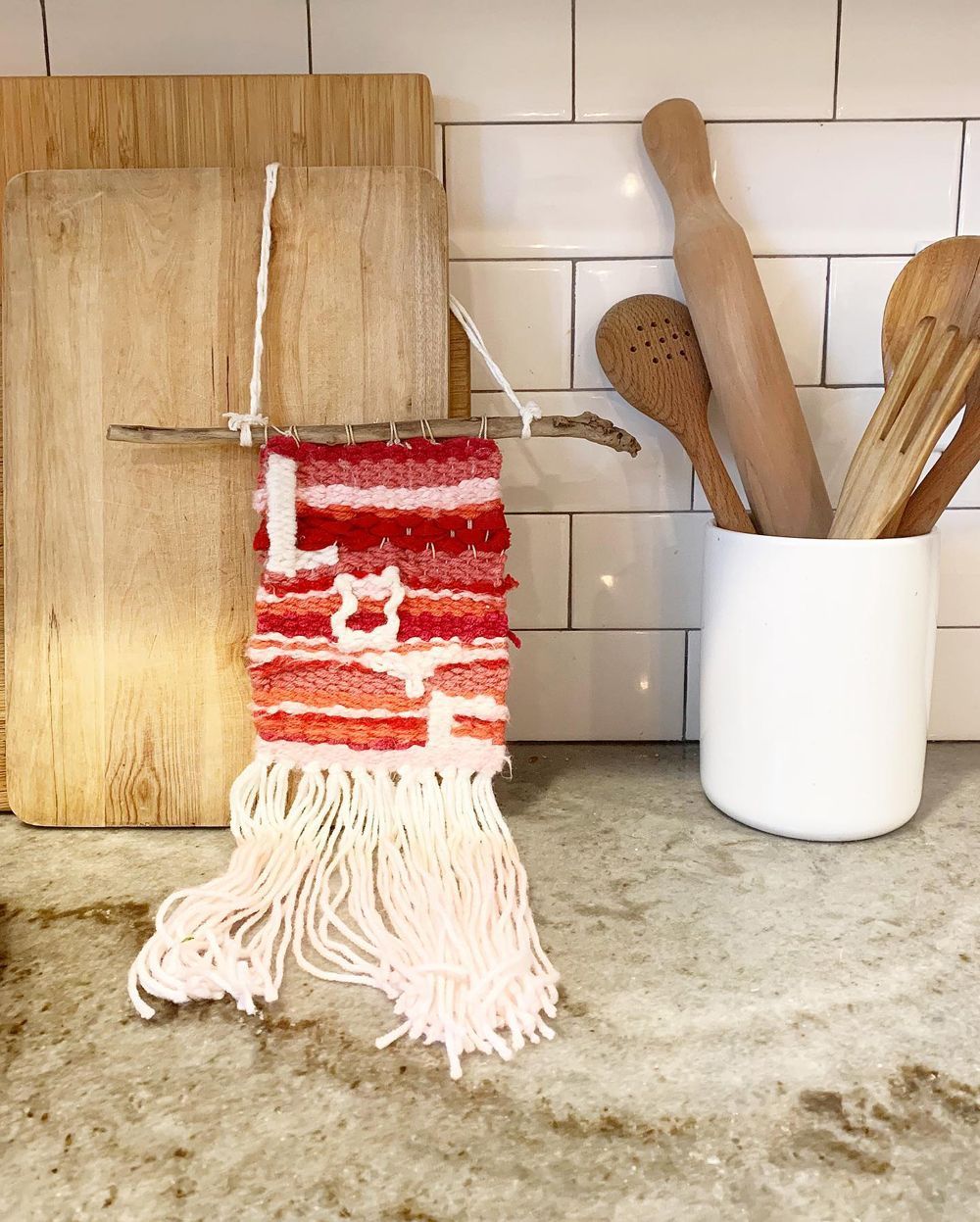 Woven fiber art with reds and whites hanging from a stick; kitchen counter with cutting boards and utensils.