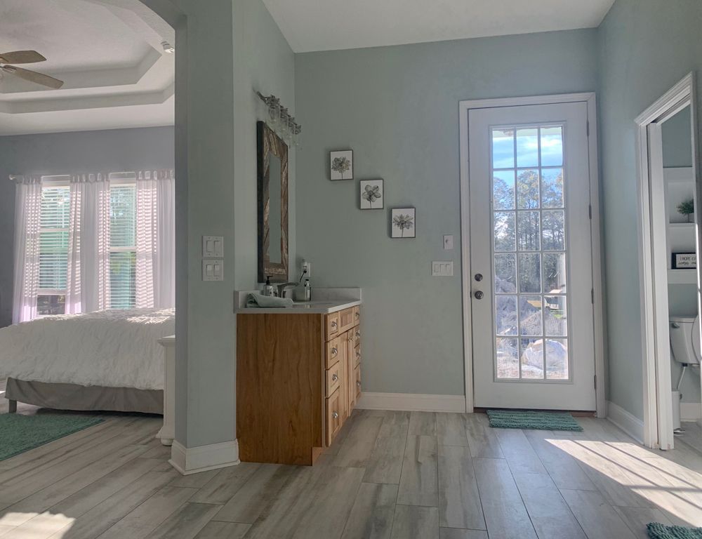 Bathroom with pale blue walls, wooden vanity, glass door, and bedroom visible through doorway.