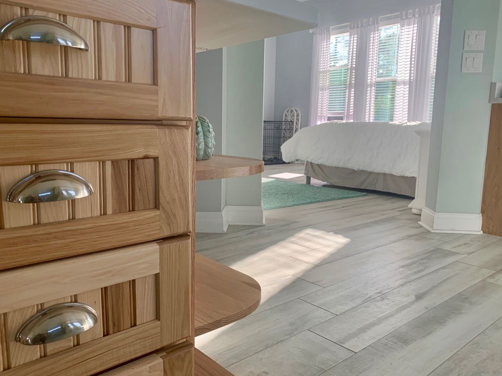 Wooden dresser in a bedroom with a bed and window. Sunlight streams across the floor.