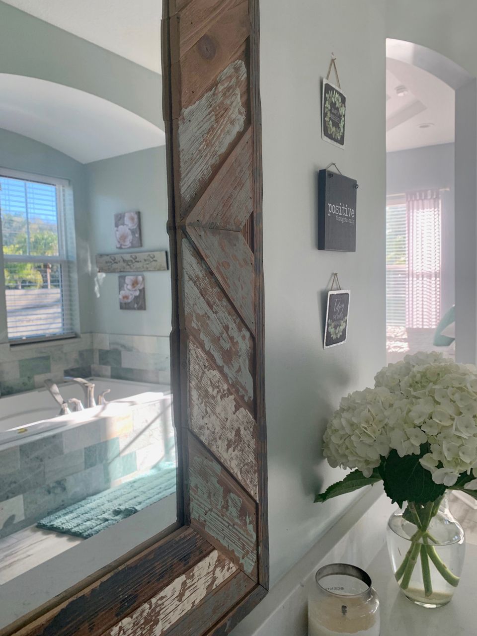 Bathroom with a large mirror framed in weathered wood. White flowers in a vase sit on a counter.