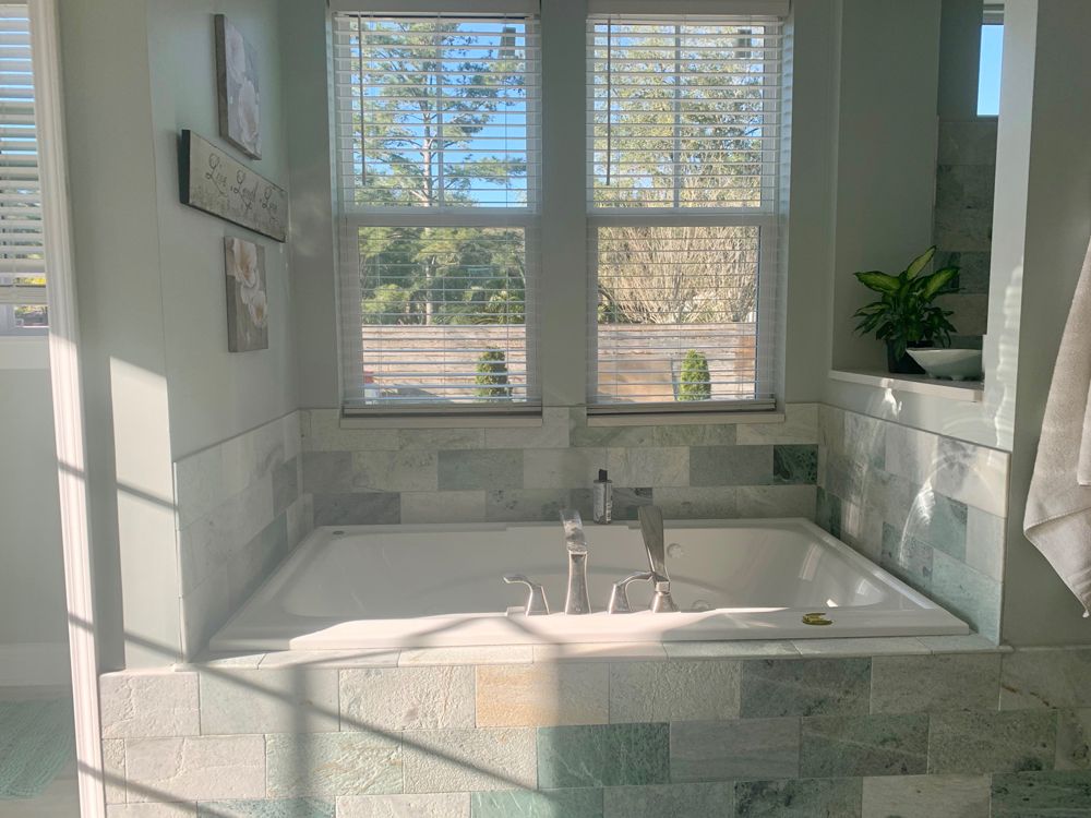 Bathroom with a built-in white tub, light green tile, two windows with blinds, and soft sunlight.