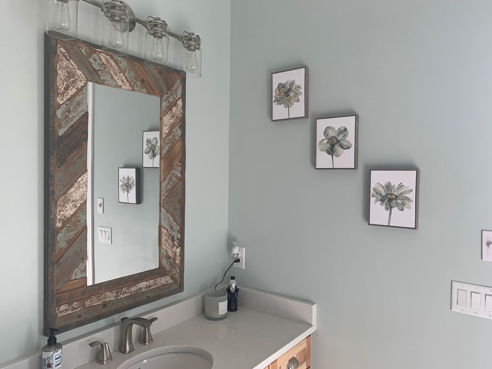 Bathroom with large rustic mirror over a sink, three flower prints on the wall, light blue walls.