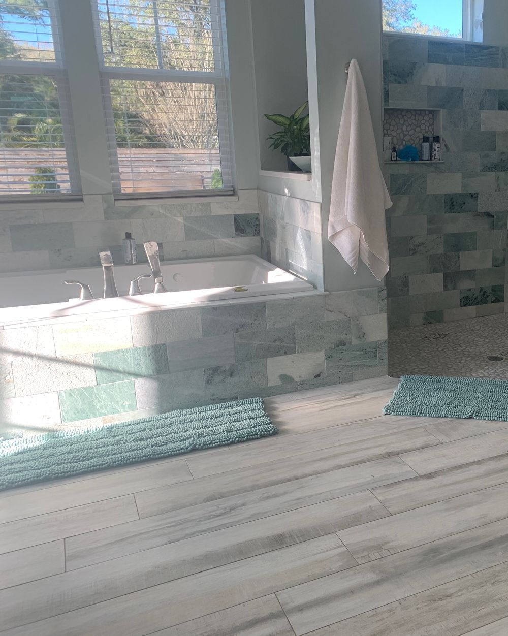 Bright bathroom with a bathtub, tiled wall, wooden floor, and a hanging towel. Blue-green accents and sunny light.