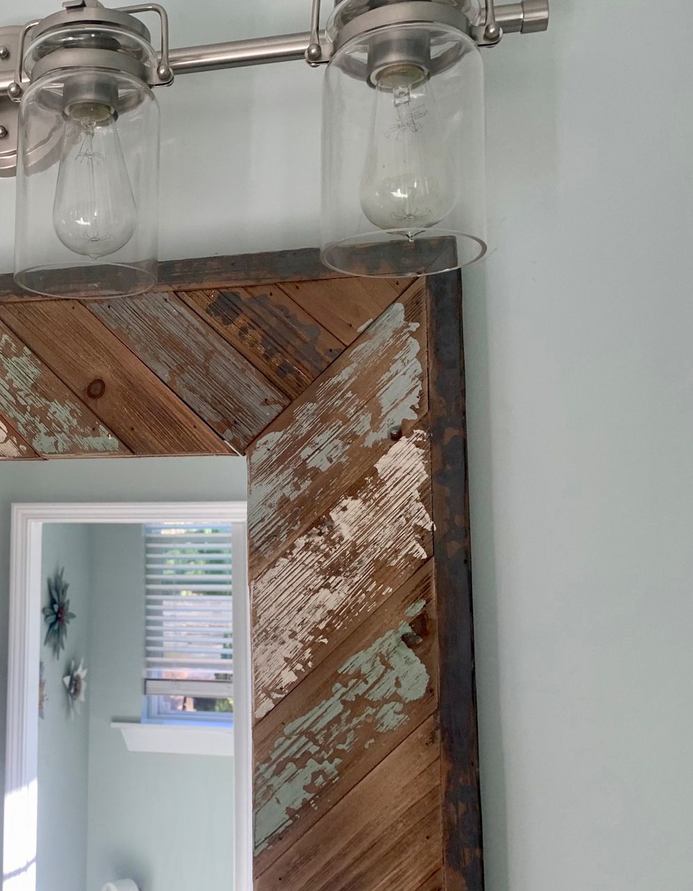 Rustic wood-framed mirror with distressed paint, mounted under a silver light fixture on a light blue wall.