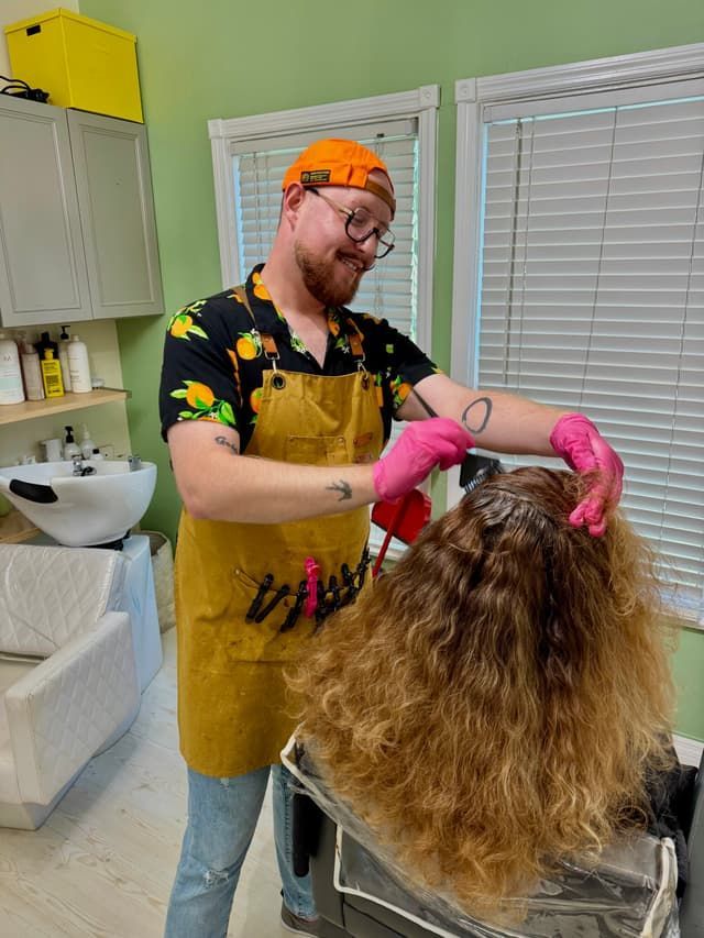 Stylist in orange cap and pink gloves grooming a client’s long hair in a salon sink
