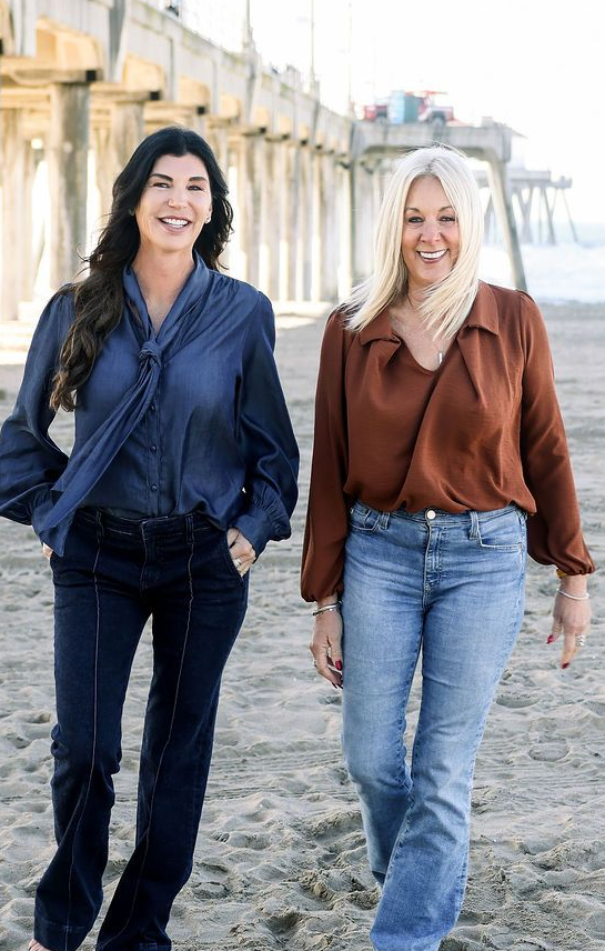 Denise Landon & Ann Stemmer Two women walking along a beach beside a pier, one in navy and one in brown, both smiling.