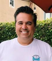 Frank Paulino Smiling man in a white T-shirt standing outdoors in front of a green hedge.