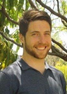 Kyle Mahoney Smiling person standing outdoors among tree branches and green leaves, wearing a dark polo shirt