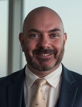 Matt Engel A smiling person wearing a dark blue suit, white collared shirt, and light-colored patterned tie.