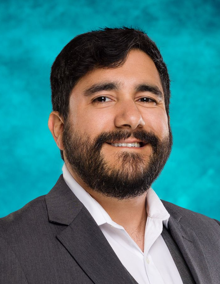 Michael Thomas Man in a gray blazer and white shirt smiling against a teal studio backdrop