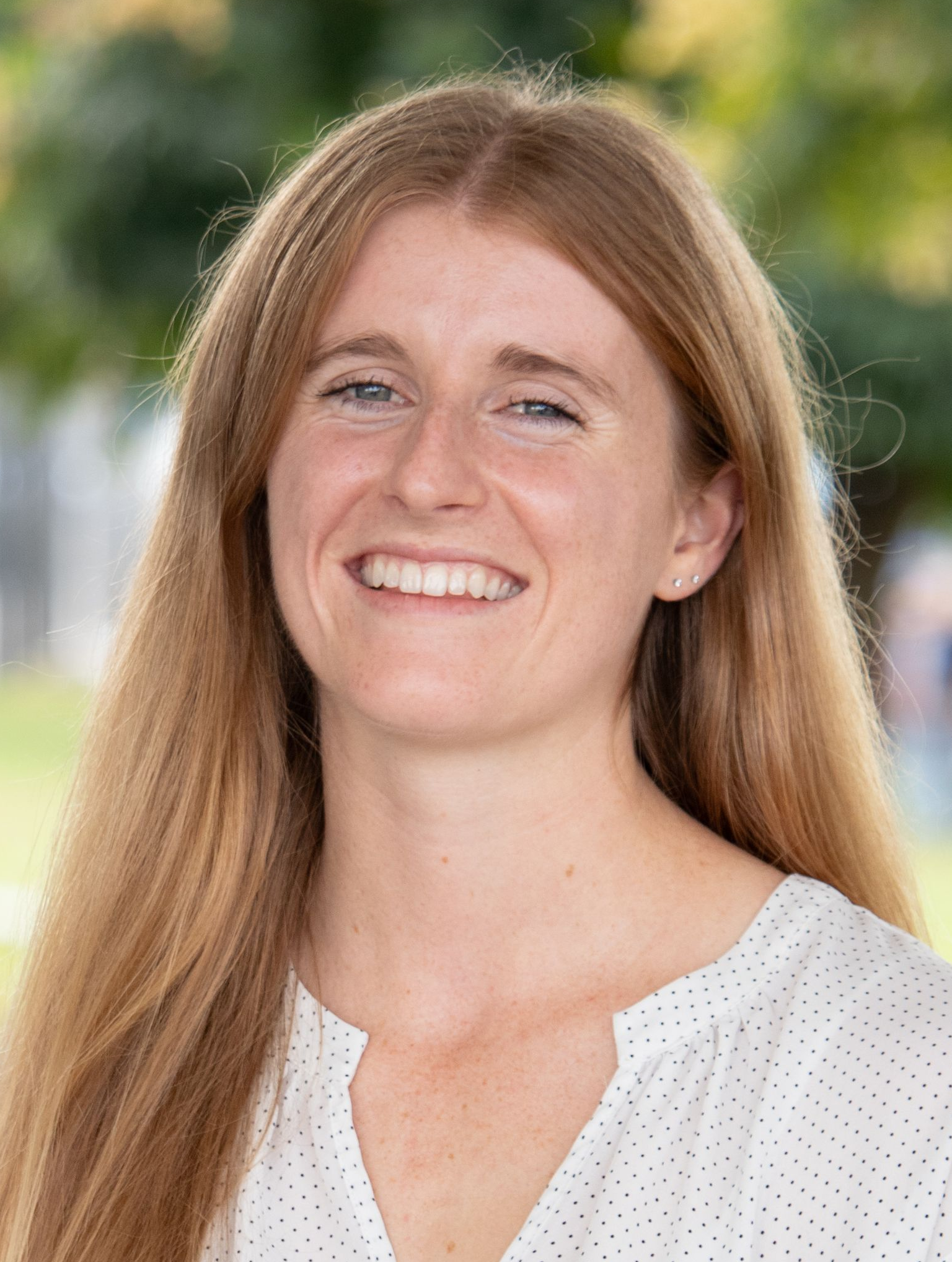 Natalie Grohmann Smiling person with long hair in a white blouse outdoors against a blurred green background. Natalie Grohmann