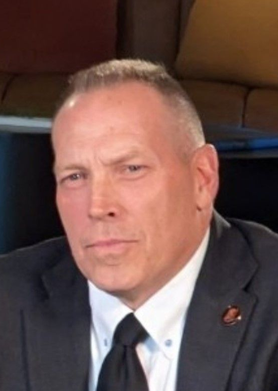 Curtis DeBerg A man with short hair wearing a dark suit, white shirt, and black tie, positioned against a blurred background.