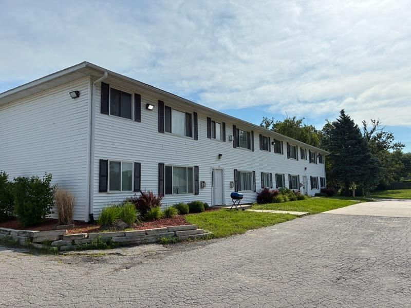Two-story white apartment building with parked car and gravel driveway under a cloudy sky
