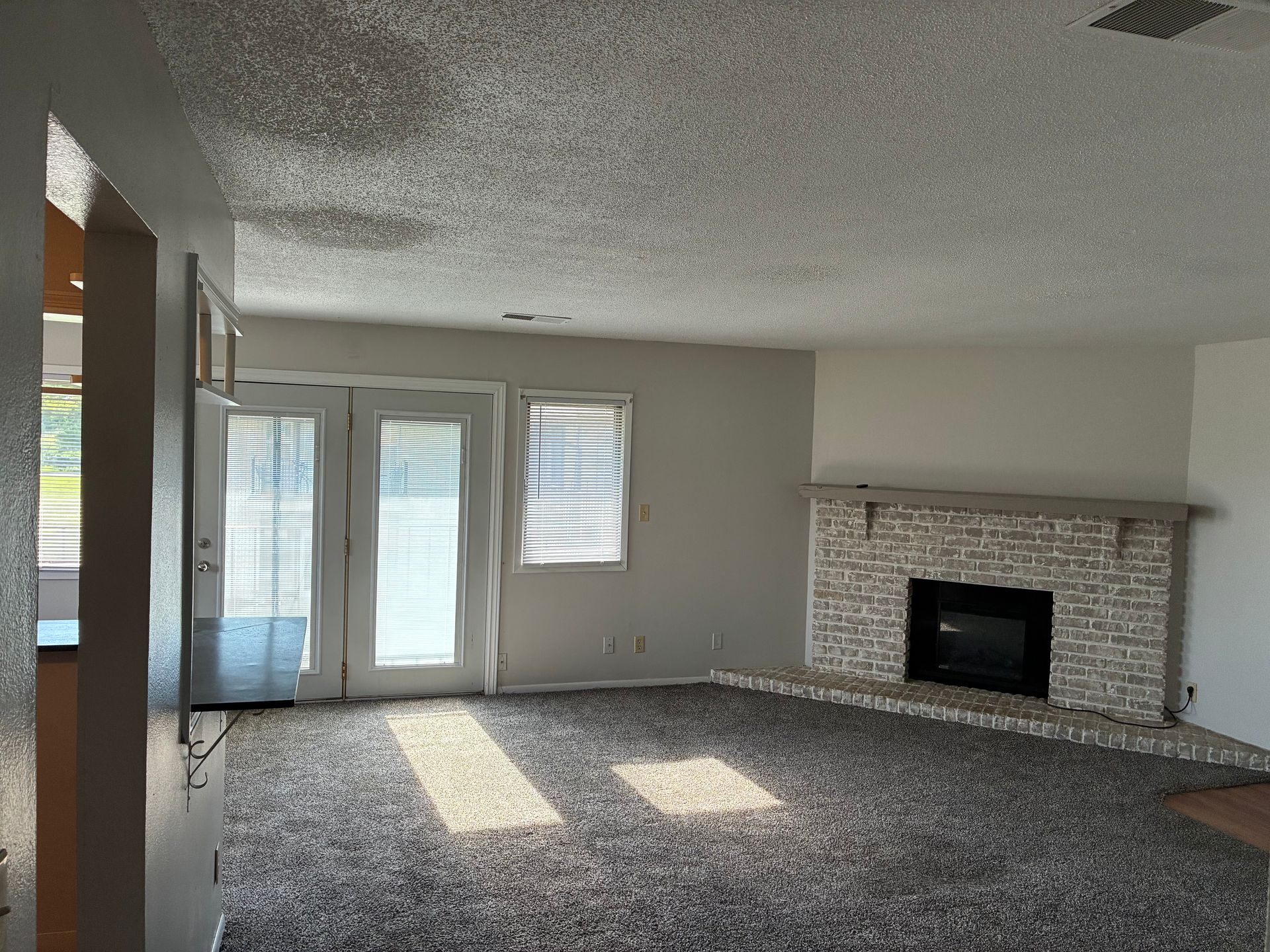 Empty living room with gray carpet, brick fireplace, and sliding glass doors with sunlight.