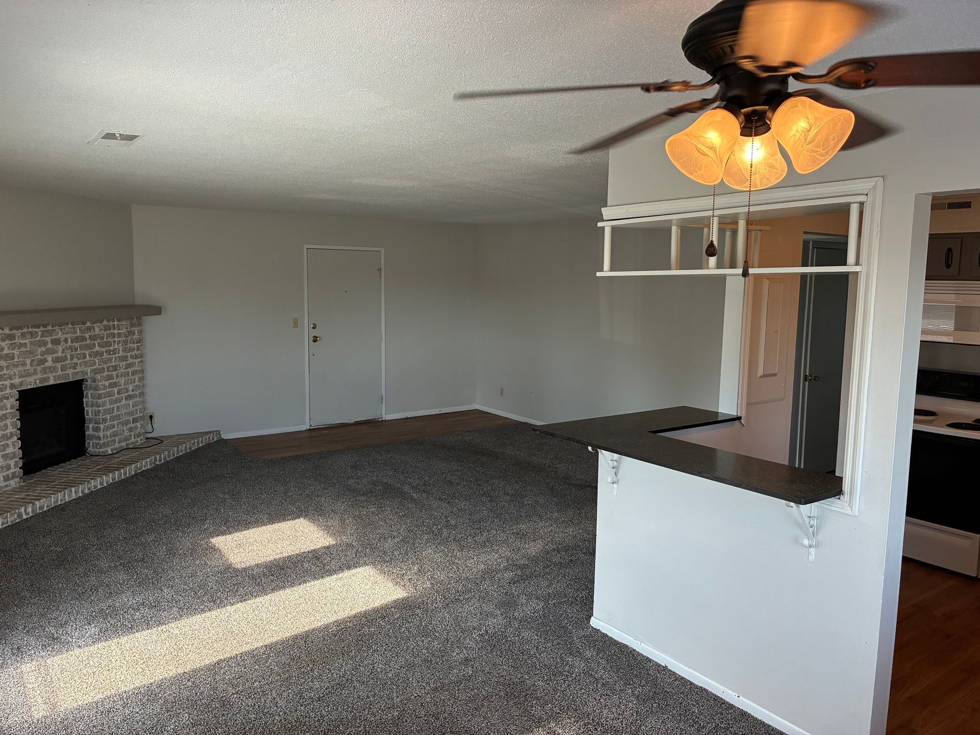 Empty carpeted living room with fireplace, ceiling fan, and pass-through counter to kitchen