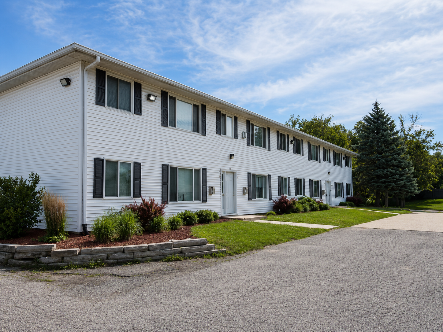 White two-story apartment building with gray shutters, landscaped front beds, and a gravel driveway
