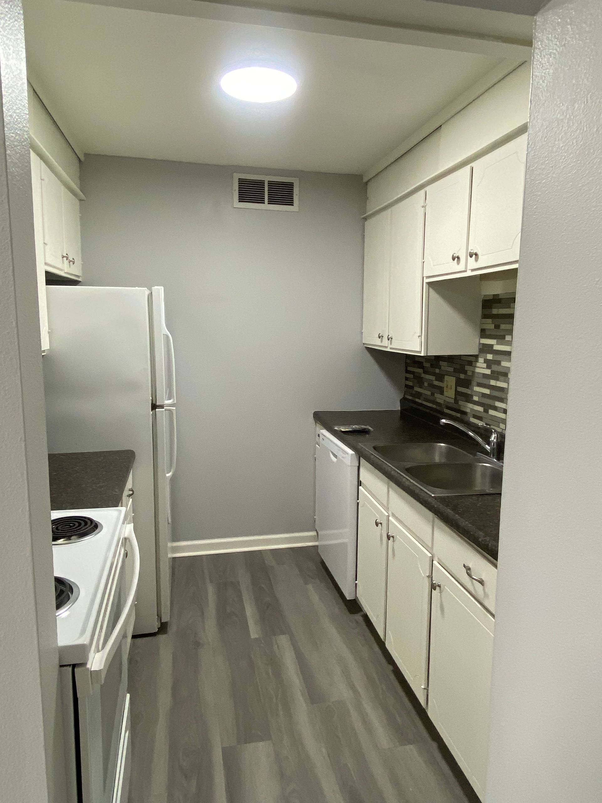 Narrow galley kitchen with white cabinets, gray floor, stove, sink, refrigerator, and overhead light.