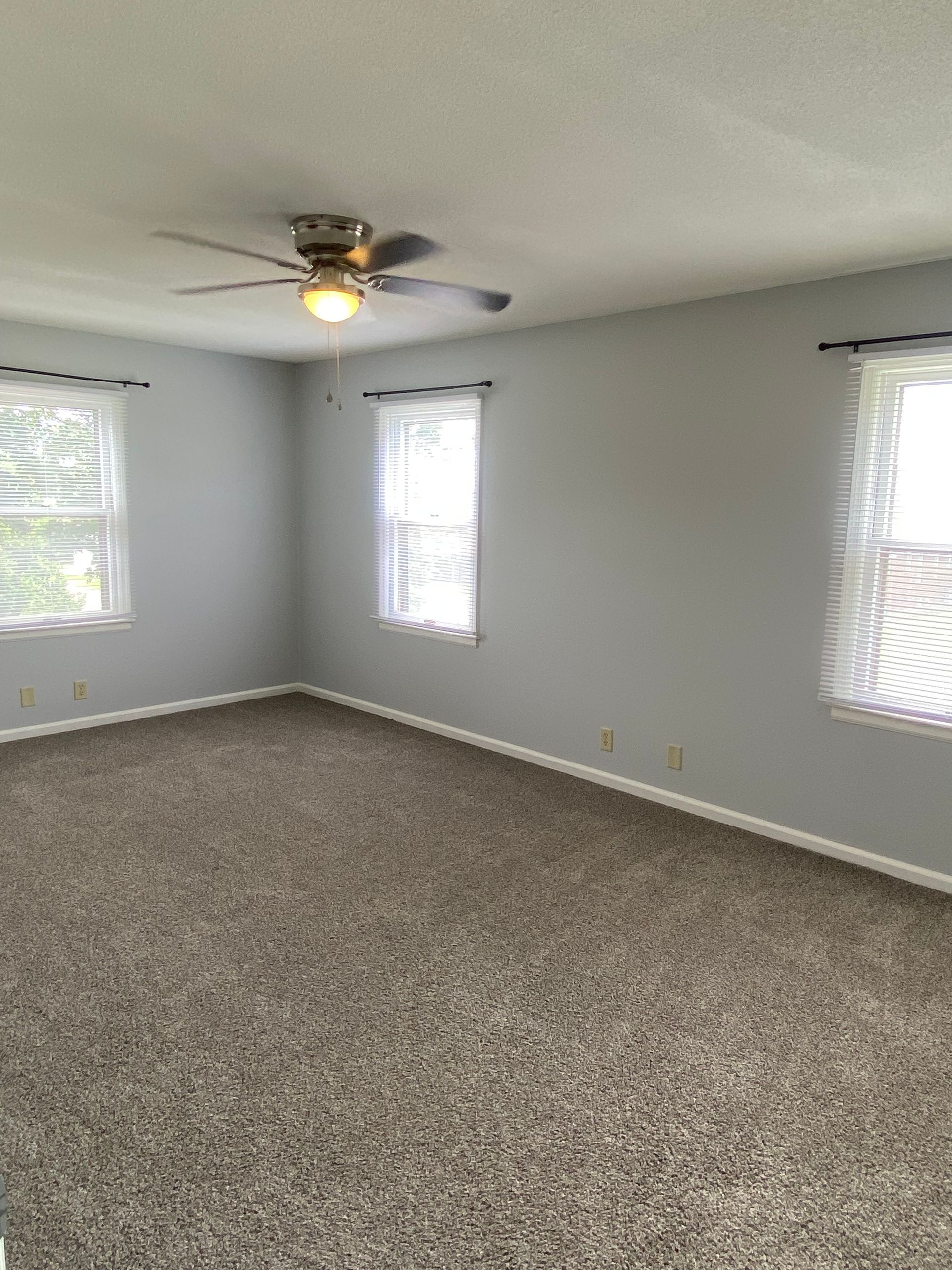 Empty carpeted room with white walls, three windows, and a ceiling fan with light on.