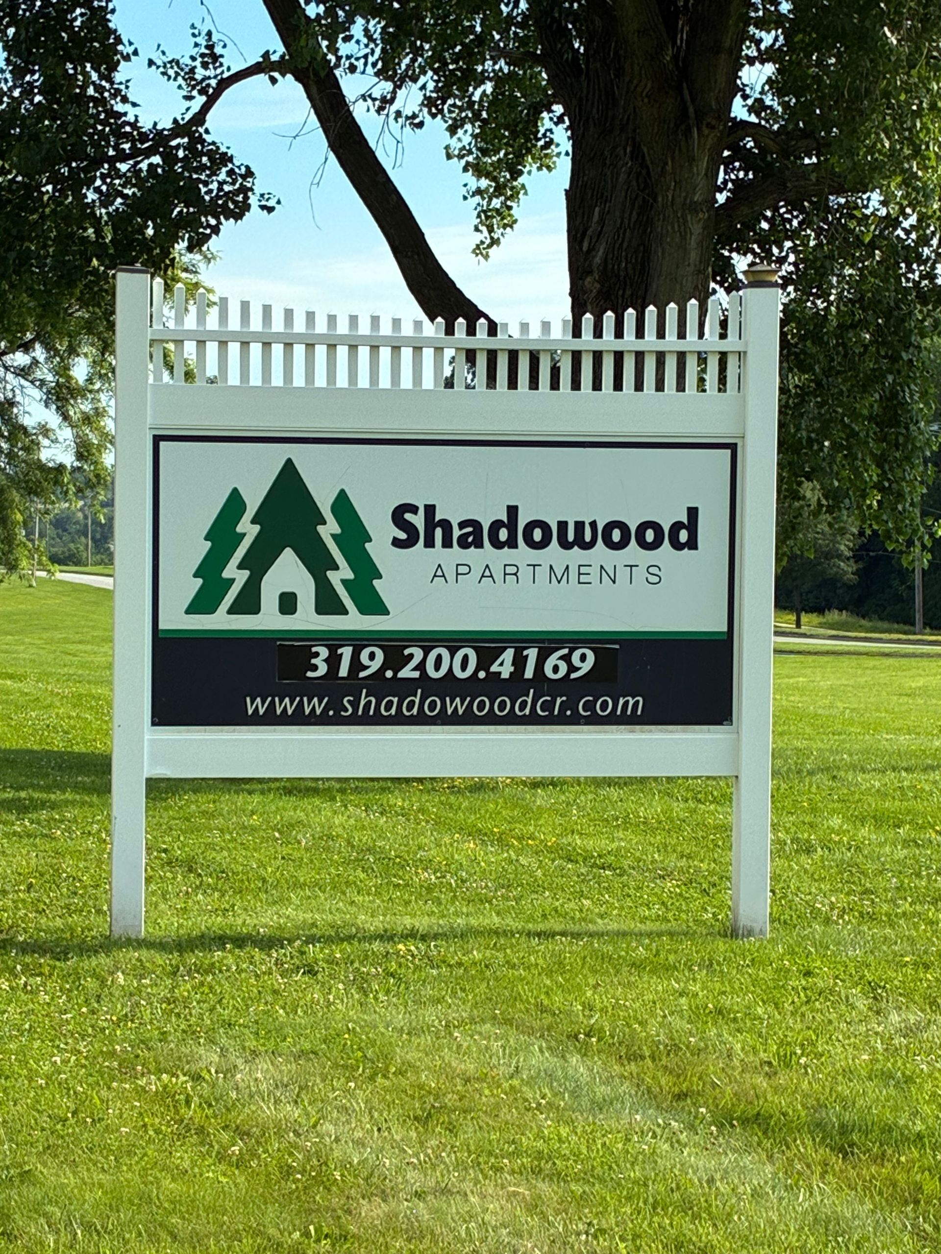 Shadowood Apartments sign on a white post in a grassy yard under trees, with phone number and website.