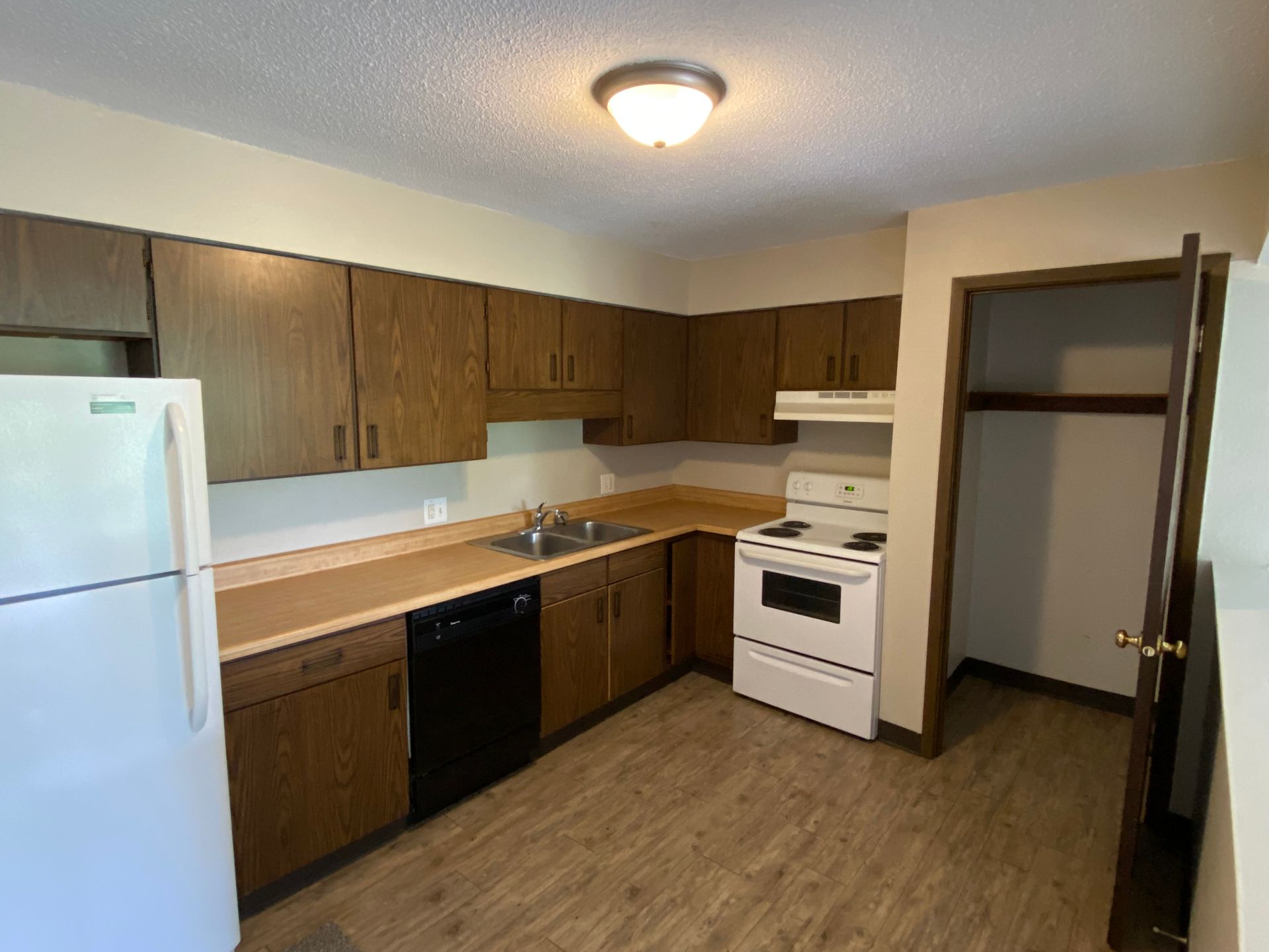 Empty kitchen with wood cabinets, white appliances, and beige counters under a ceiling light.