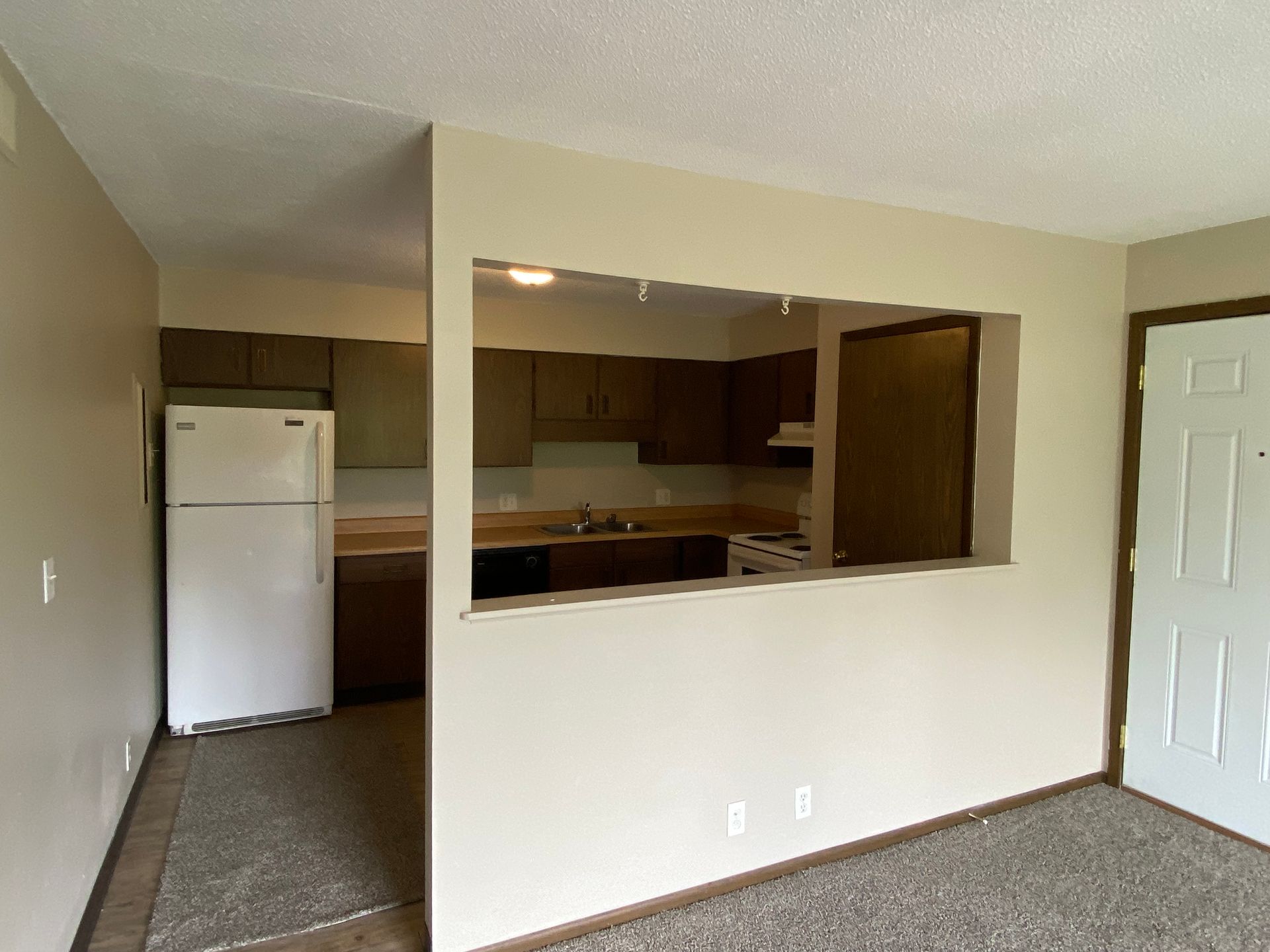 Empty apartment living area with pass-through to kitchen, white fridge, beige walls, and carpeted floor