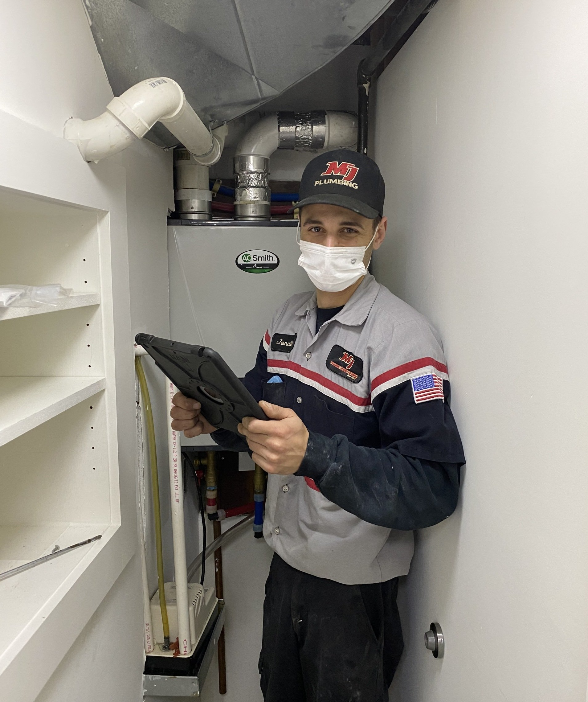 Technician in a narrow utility closet holding a tool, with exposed pipes overhead and shelves nearby