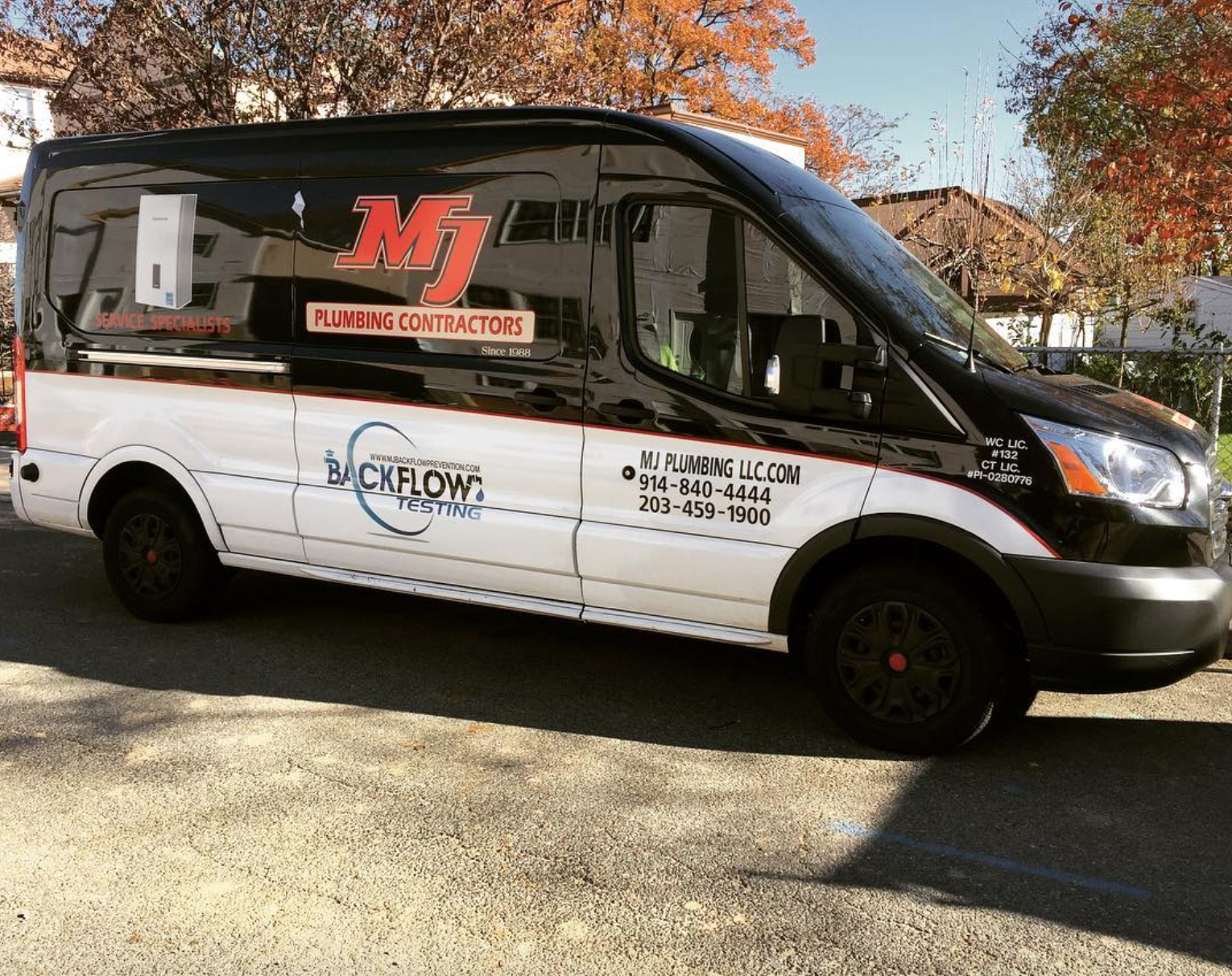White-and-black van with M&M Plumbing Contractor logo parked outdoors in autumn.