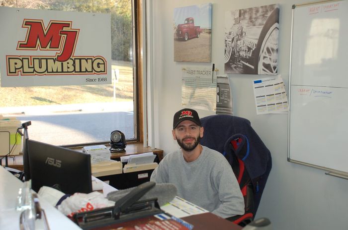 Man seated at a desk in a plumbing office with computer monitors, whiteboard, and MJ Plumbing signage.