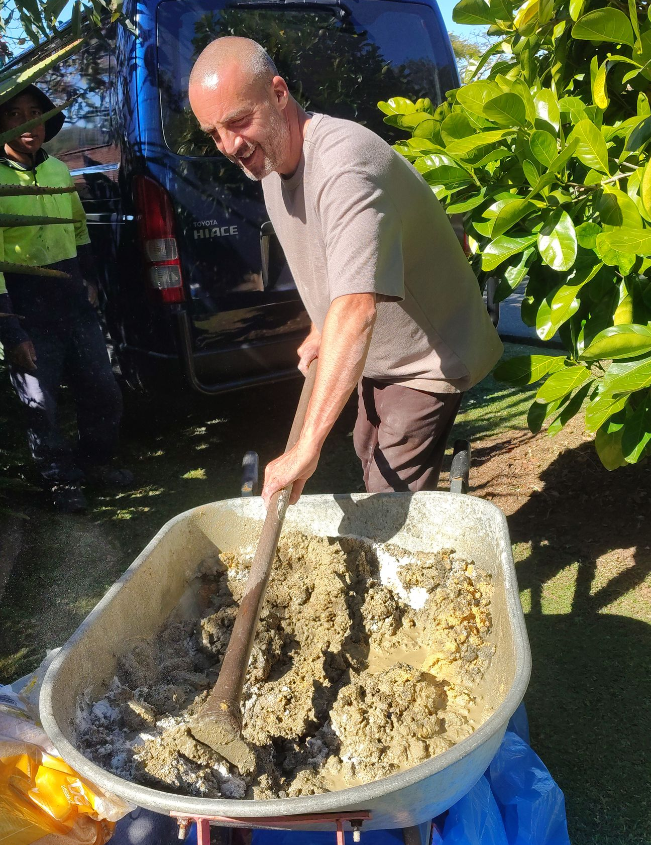 Man mixing slury for roof repointing in a large basin outdoors beside leafy plants and a parked vehicle