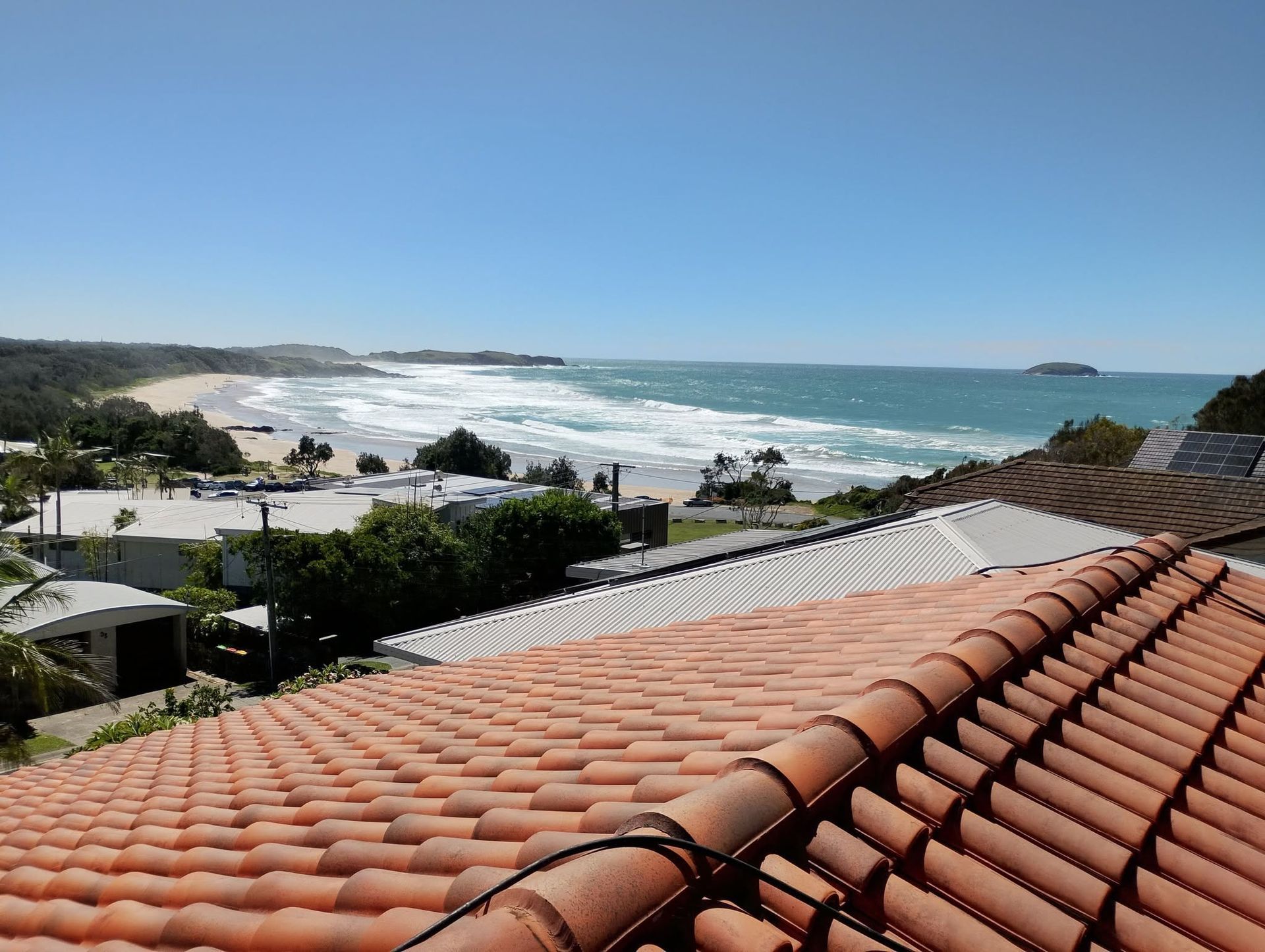 Coffs Coastal neighbourhood with red-tiled roof, ocean waves, and a sunny blue sky