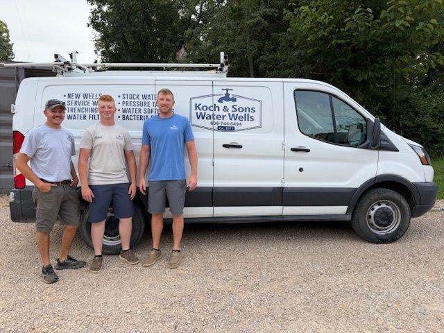 Three workers stand beside a white service van with a company logo on a gravel driveway.