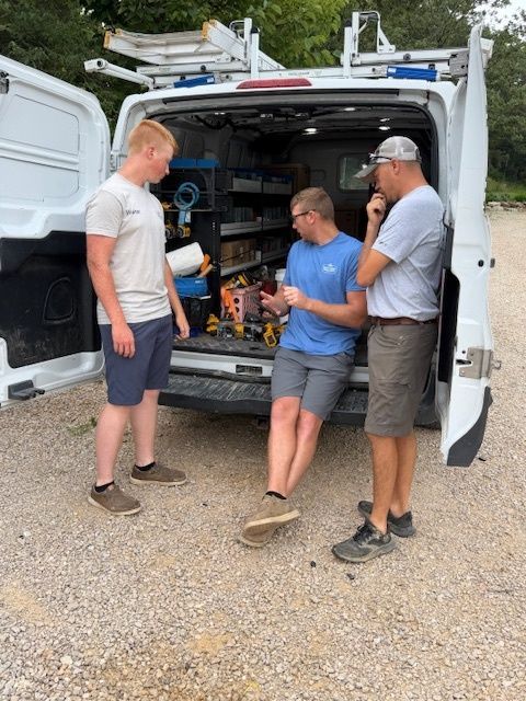 Three men chat at the open back of a white van packed with tools and supplies.