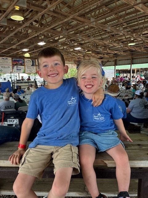 Two smiling children in blue shirts sit on a bench in a crowded pavilion.