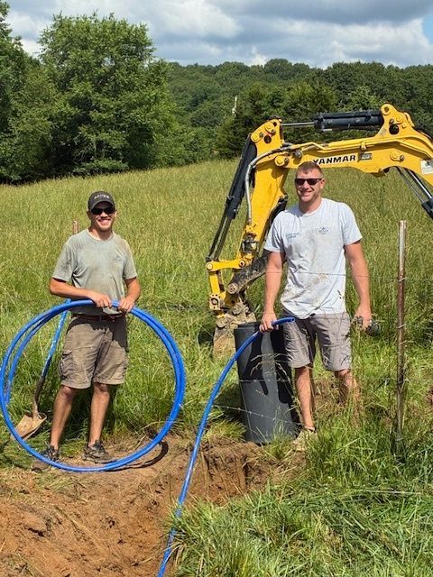 Two workers beside a yellow excavator, holding blue tubing in a grassy field.