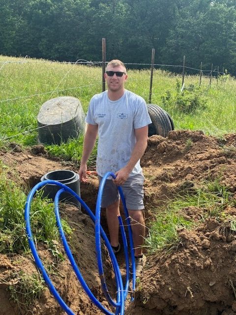 Man standing in a dirt trench holding blue hoses in a grassy field with large concrete pipes nearby