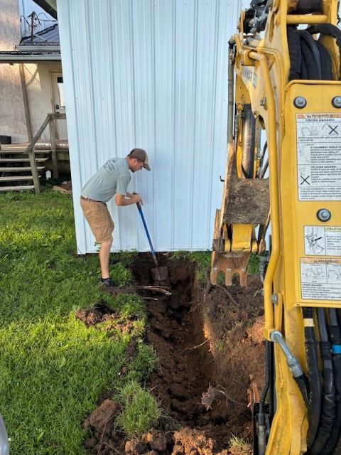 Worker digging a trench beside a blue building with a yellow excavator nearby